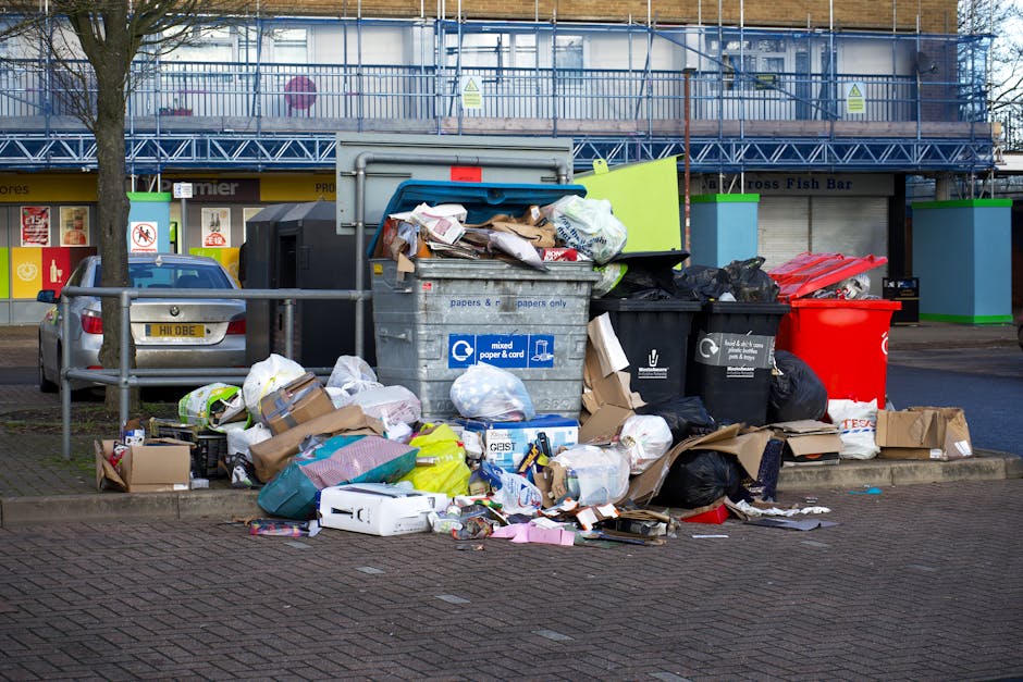 A large pile of mixed waste materials arranged outside a commercial building on a paved area, including overflowing rubbish bins and scattered trash. The waste consists of cardboard boxes, paper bags, plastic bottles, black and black-and-white plastic garbage bags, and various packaging materials, some of which are crumpled or torn. A prominent grey recycling bin labeled for mixed paper and card is partially open with papers protruding from the top, alongside several black bins with lids, one black bin with a partially open lid, and a bright red bin to the right. The debris extends onto the surrounding pavement, with discarded boxes, plastic packaging, and other refuse scattered across the ground. Behind the waste collection area, the background features a modern building with a blue metal scaffolding structure and a storefront with signage, while a silver car is parked nearby, partially obscured by a small metal railing. The scene suggests an instance of waste overflow that a rubbish removal service like Waste Removal Uxbridge could professionally address, ensuring proper disposal and site clearance within an urban environment.