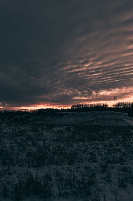 A landscape scene featuring a snow-covered hill in the foreground, with patches of bare shrubbery visible beneath a dark, cloudy sky illuminated by a faint orange glow near the horizon. In the distance, there are silhouettes of leafless trees and a utility pole, indicating a rural or semi-rural environment. The overcast sky has textured, rippled cloud formations, creating a moody and dramatic atmosphere. The image's muted tones and natural lighting evoke an outdoor setting that might be associated with waste management or site clearance activities, reflecting a neutral, observational focus suitable for a rubbish removal service page.