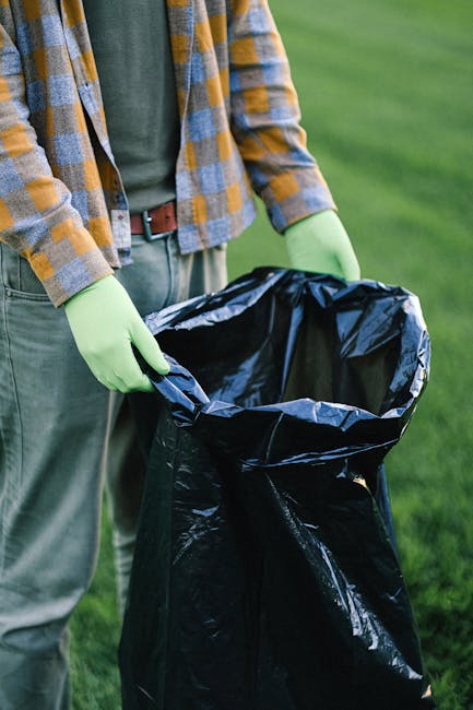 A large pile of mixed waste materials arranged outside a commercial building on a paved area, including overflowing rubbish bins and scattered trash. The waste consists of cardboard boxes, paper bags, plastic bottles, black and black-and-white plastic garbage bags, and various packaging materials, some of which are crumpled or torn. A prominent grey recycling bin labeled for mixed paper and card is partially open with papers protruding from the top, alongside several black bins with lids, one black bin with a partially open lid, and a bright red bin to the right. The debris extends onto the surrounding pavement, with discarded boxes, plastic packaging, and other refuse scattered across the ground. Behind the waste collection area, the background features a modern building with a blue metal scaffolding structure and a storefront with signage, while a silver car is parked nearby, partially obscured by a small metal railing. The scene suggests an instance of waste overflow that a rubbish removal service like Waste Removal Uxbridge could professionally address, ensuring proper disposal and site clearance within an urban environment.