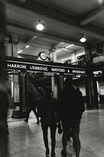 A black and white photograph taken inside a train station shows two silhouetted figures walking towards the camera, with one carrying a bag in their right hand. Above them, a large illuminated sign displays the destinations Harrow, Uxbridge, Watford, and Amersham in bold, uppercase letters. In the background, a staircase leading upward is visible, flanked by brick columns and metal railings, while the station's ceiling features exposed beams and several round ceiling lights. To the left of the stairway, a clock mounted on the wall indicates the time as approximately 8:20. The tiled floor reflects the station’s lighting, creating a clean, functional atmosphere suitable for rail travellers. The image’s overall tone emphasizes the everyday activity within a busy railway hub, aligning with the context of providing efficient, alternative waste handling or rubbish removal services in public transportation environments, as provided by Waste Removal Uxbridge.