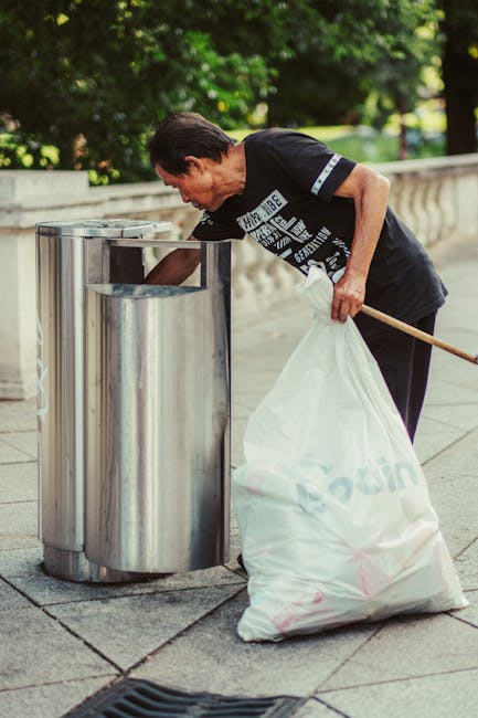 A man with short dark hair, wearing a black T-shirt with white text and black trousers, is depicted disposing of rubbish into a tall, cylindrical stainless steel trash bin on an outdoor paved walkway. He is bent slightly forward, holding a long wooden stick in his left hand and a large white sack filled with waste in his right hand, which is partially supported by his hip. The stainless steel bin features a wide rectangular opening at the top, with a smooth, reflective surface that shows slight distortions from contact. The background includes lush green trees and a stone balustrade, indicating a park or garden setting. The scene appears to be during daylight with natural lighting illuminating the area evenly. The image visually aligns with services like waste removal or rubbish collection, emphasizing private waste disposal methods such as on-site clearance, consistent with offerings by Waste Removal Uxbridge.